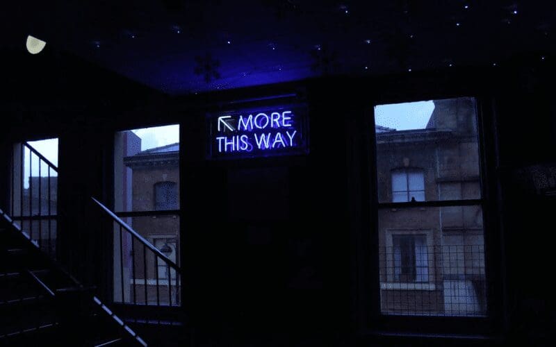Stairwell with a blue glowing neon sign saying 'MORE THIS WAY'. Indie Wire