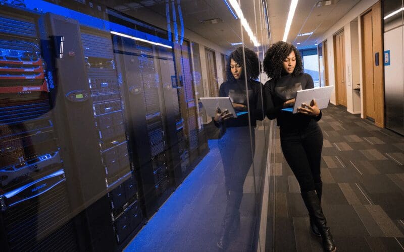 Girl standing next to server room working on her laptop.