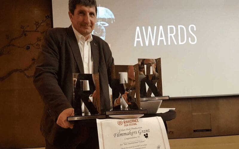 A director standing in front of a presentation with his awards at a Raindance film festival.